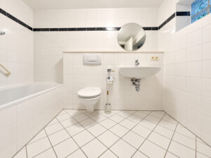 Weiß gefliestes Badezimmer mit Badewanne, Toilette und rundem Spiegel